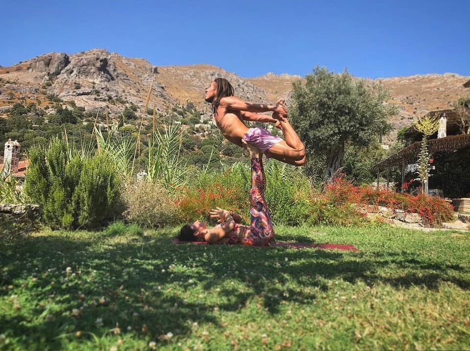 Karakaya Retreat'in dağ manzaralı bahçesinde acro-yoga: sırt üstü yatan base ayaklarıyla üstteki kişiyi havada taşıyor, üstteki kişi bir ayağını iki eliyle tutuyor