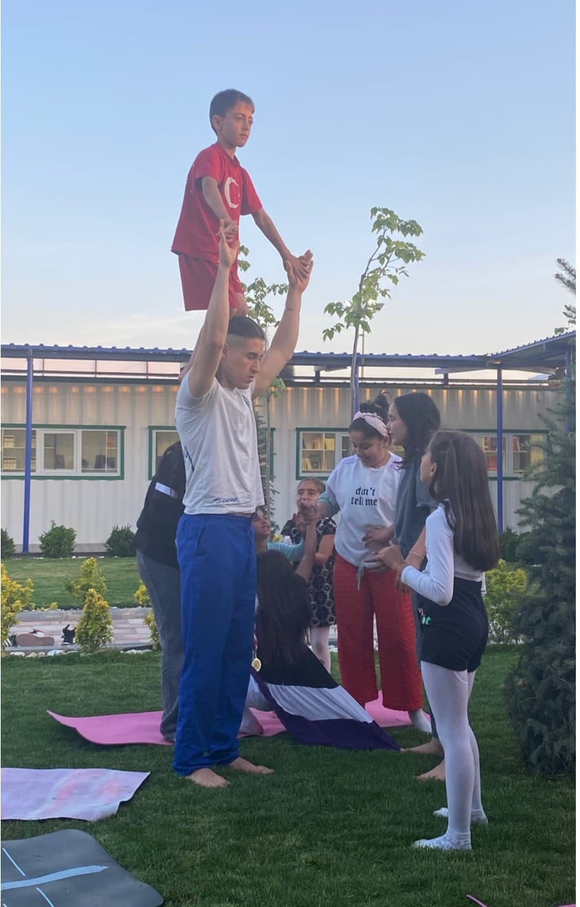 Türker çimlerde bir çocuğu ellerinin üstünde tutarken, çevresinde acro-yoga yapan çocuklar
